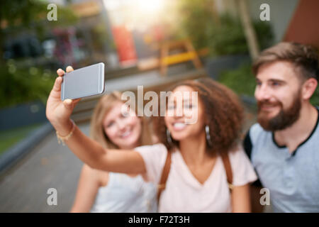 Outdoor shot of young woman taking self portrait on mobile phone with her friends. Three young friends taking a selfie with smar Stock Photo