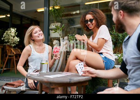 Three friends sitting in outdoor cafe and playing cards and having fun. Happy young people at sidewalk coffee shop enjoying play Stock Photo