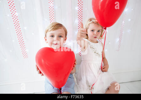 Young boy and girl posing for a picture in a photographers studio, holding red balloons.