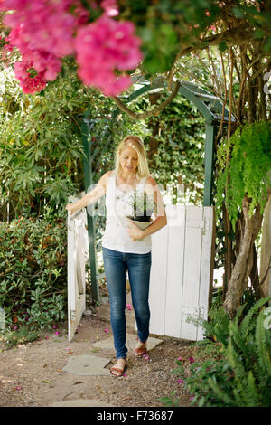Blond woman entering a garden through a white wooden gate, carrying a plant pot. Stock Photo