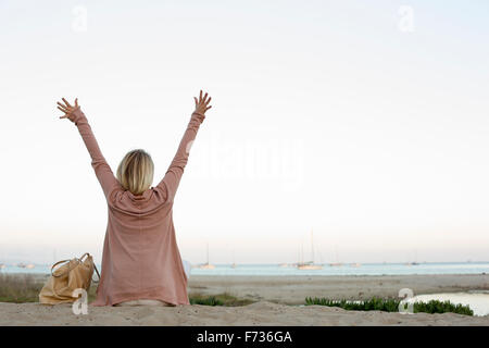 Blond woman sitting on a sandy beach, arms raised. Stock Photo