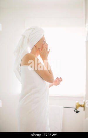 Woman wrapped in a white towel standing in a bathroom, applying cream to her face. Stock Photo