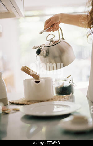 Woman standing in a kitchen pouring hot water from a kettle into a tea pot. Stock Photo