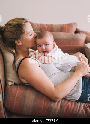 A woman lying on a sofa playing with a baby girl, kissing her head. Stock Photo