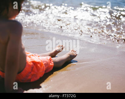 A boy sitting on the sand at the water's edge watching the waves reach the beach. Stock Photo