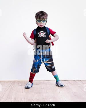 A boy standing with his legs apart posing in fancy dress, wearing a pirate tee-shirt, eye goggles and long shorts. Stock Photo