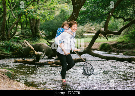 A woman giving a boy a piggyback and holding a shrimping net wading in a shallow stream. Stock Photo