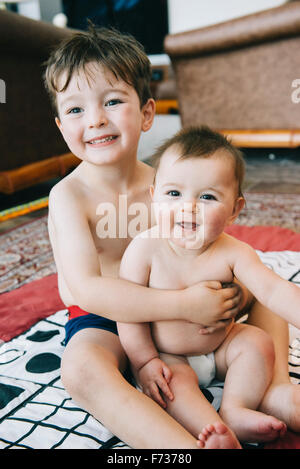 Two siblings, a boy and his baby sister sitting on a rug laughing. Stock Photo