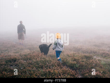An adult and a child with a dog, walking through heather in autumn mist.