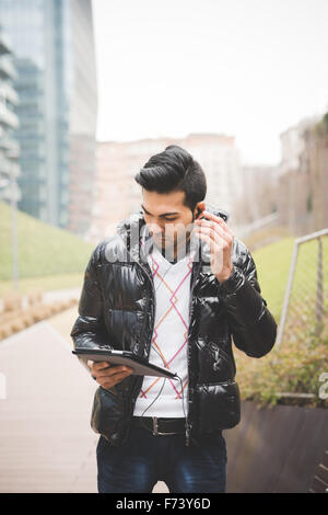 Knee figure of a young handsome indian businessman listening music with earphones while working on a tablet connected online looking downward on the screen - business, work, study, technology concept Stock Photo