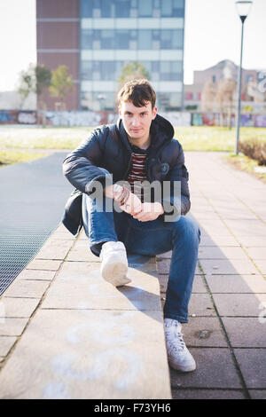 young handsome caucasian brown hair man sitting on a small wall, looking in camera, smiling - youth, carefree concept Stock Photo