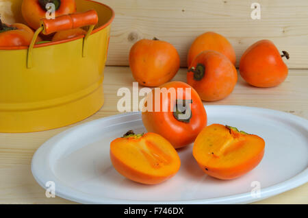 Kaki apples on white plate and orange tin bowl with fruits in background Stock Photo