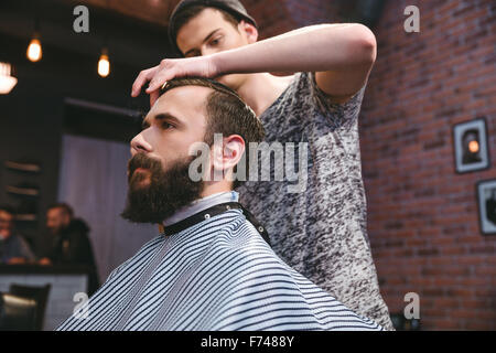 Young handsome barber cutting hair of young attractive bearded hipster in barbershop Stock Photo