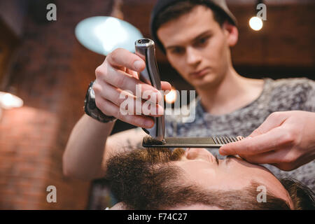 Young concentrated skillful barber making perfect beard to handsome bearded man using trimmer and comb in hair salon Stock Photo