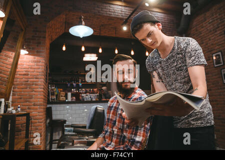 Young hairstylist and attractive bearded client looking through magazine sitting in barbershop Stock Photo