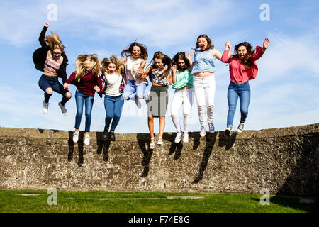 Eight teenage girls having fun holding hands and jumping off the Broughty Ferry Castle wall on a sunny day in Dundee, UK Stock Photo