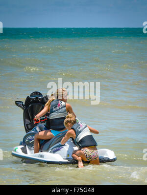 Young woman in bikini bottoms and life jacket struggles to crawl onto the back of a jet ski with blond woman driving blue waves Stock Photo