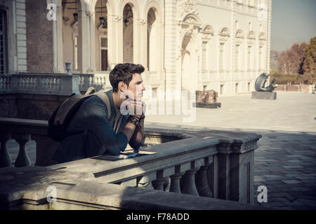 Half Body Shot of a Thoughtful Handsome Young Man, a Tourist, Holding a Guide, Looking Away Outside Historic Building in Europea Stock Photo