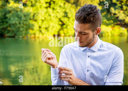 Portrait of contemplative light brown haired young man wearing white shirt beside picturesque river or lake, looking at shirt cu Stock Photo