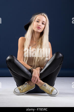 Attractive blond woman sitting crossing legs in leather jeans with a black cap Stock Photo