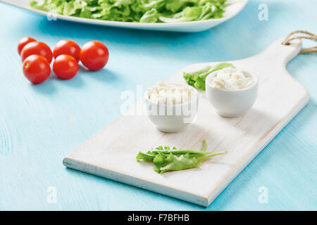 Cottage cheese in two white bowls on blue wooden table, salad and tomato Stock Photo