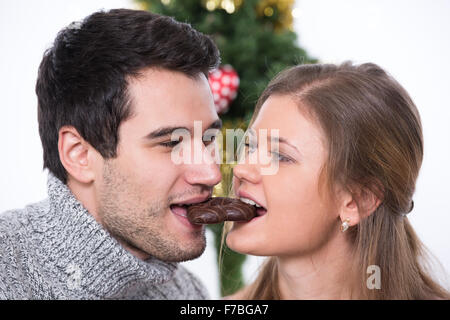 young couple, man and woman, in front of Christmas tree, both eating one cake, white background Stock Photo