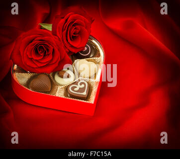 Red roses and chocolate pralines in golden heart shaped gift box on satin background