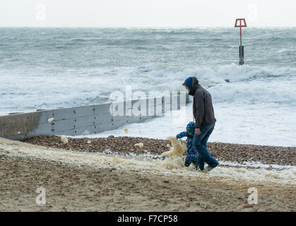 29th November 2015, UK weather, Boscombe, Bournemouth. Man and child walking on the beach in coats with foam from the sea blowing across in the strong wind of a winter storm.