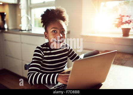 Black girl sitting playing on a laptop computer at home looking at the camera with a joyful expression of amazement and wonder Stock Photo