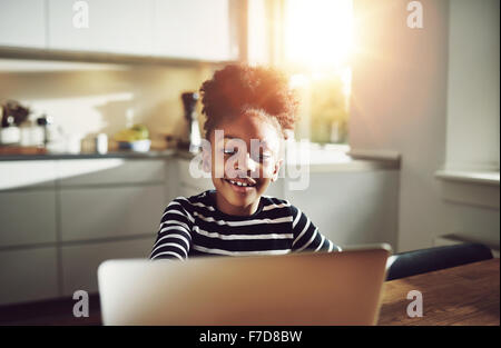 Cute young black girl playing on a laptop computer smiling happily as she surfs the internet while relaxing at a table at home,