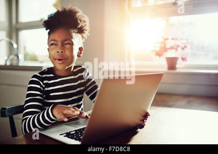 Fun little black girl with her frizzy afro hair up on top of her head sitting at a table at home working on a laptop computer sc Stock Photo