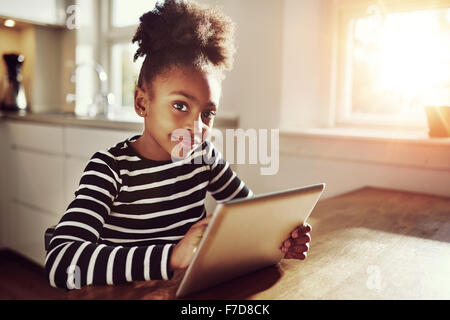 Thoughtful young black girl sitting watching the camera with a pensive expression as she browses the internet on a tablet comput Stock Photo