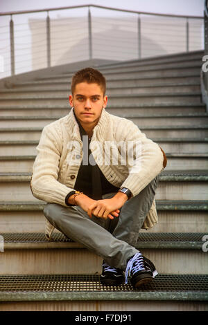 Well dressed handsome young man in casual clothes sitting cross legged on the lower part of a stairway outdoor in winter Stock Photo