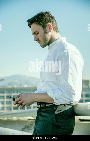 Handsome trendy man wearing white shirt standing and looking down at a cell phone that he is holding, outdoor in city setting Stock Photo