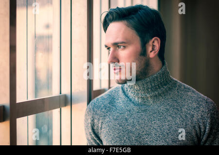 Handsome serious man standing inside modern building next to big window, wearing wool sweater,  looking out Stock Photo