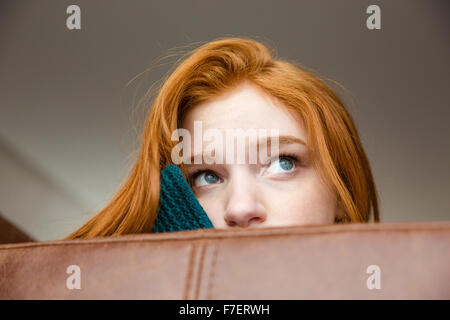 Unconfident shy redhead girl peeping over brown leather sofa and looking away Stock Photo