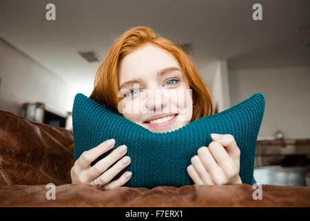 Happy attractive smiling redhead girl lying on brown leather sofa and hugging knitted pillow Stock Photo