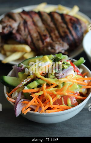 A bowl of Greek salad in a white bowl with steak and chips in the background Stock Photo