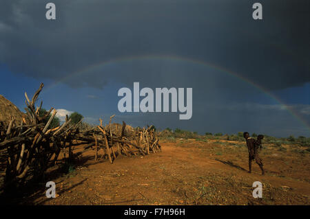 A double rainbow arches over a Hamar village and a Hamar girl who carries and infant on her hip, in South Omo, Ethiopia Stock Photo