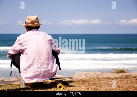 Man watching the sea and the waves from the shore sitting on his skateboard Stock Photo