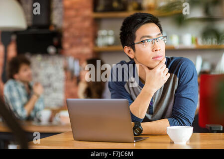 Thoughtful handsome young asian man in glasses sitting with laptop in cafe and looking away Stock Photo