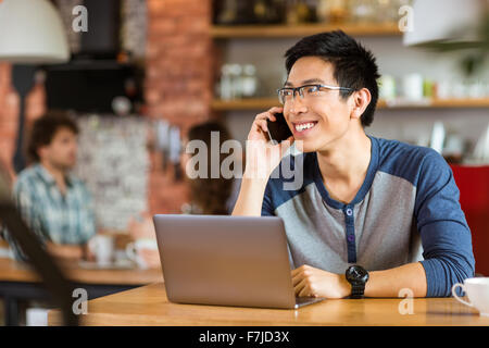 Positive attractive asian man in glasses using laptop and talking on cellphone in cafe Stock Photo