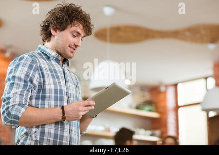 Attractive thoughtful smiling curly young male in plaid shirt using tablet at home Stock Photo