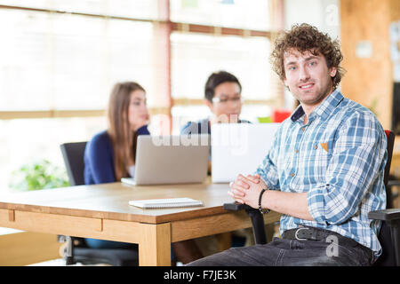 Smiling happy young curly male in checkered shirt working in team at wooden table and writing into notebook Stock Photo