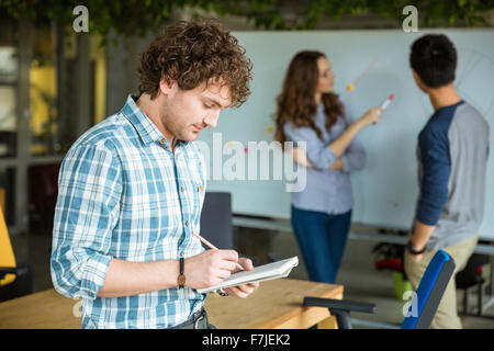 Concentrated pensive young curly male in checkered shirt writing in notebook and working in team with partners Stock Photo