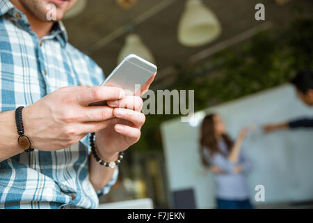 Closeup of hands of young man in checkered shirt using mobile phone while his partners arguing Stock Photo