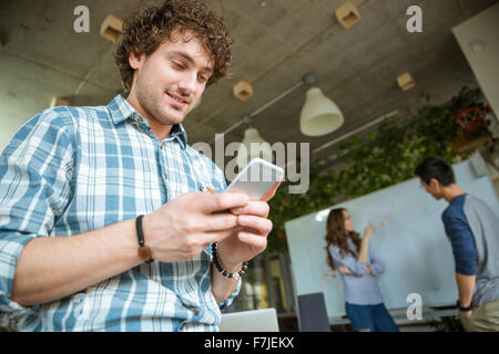 Smiling young curly man in plaid shirt using cellphone while his friends studying and making homework Stock Photo