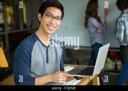 Happy smiling young asian man in glasses holding laptop and studying with friends in classroom Stock Photo