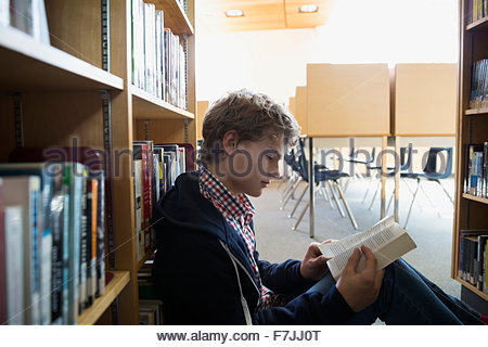 High school student reading book on library floor
