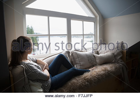 Pensive young woman looking out window lake view